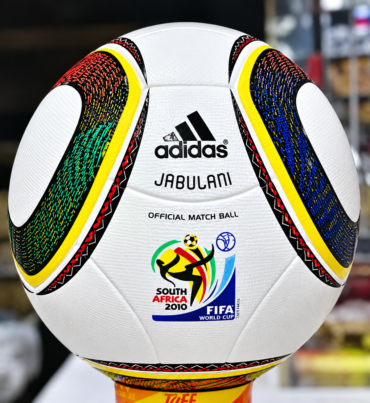 A white Adidas Jabulani soccer ball with colorful curved panels, displayed on a stand. The ball is labeled “adidas,” “JABULANI,” “OFFICIAL MATCH BALL,” and features the South Africa 2010 FIFA World Cup logo.