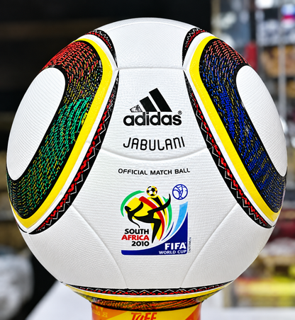 A white Adidas Jabulani soccer ball with colorful curved panels, displayed on a stand. The ball is labeled “adidas,” “JABULANI,” “OFFICIAL MATCH BALL,” and features the South Africa 2010 FIFA World Cup logo.