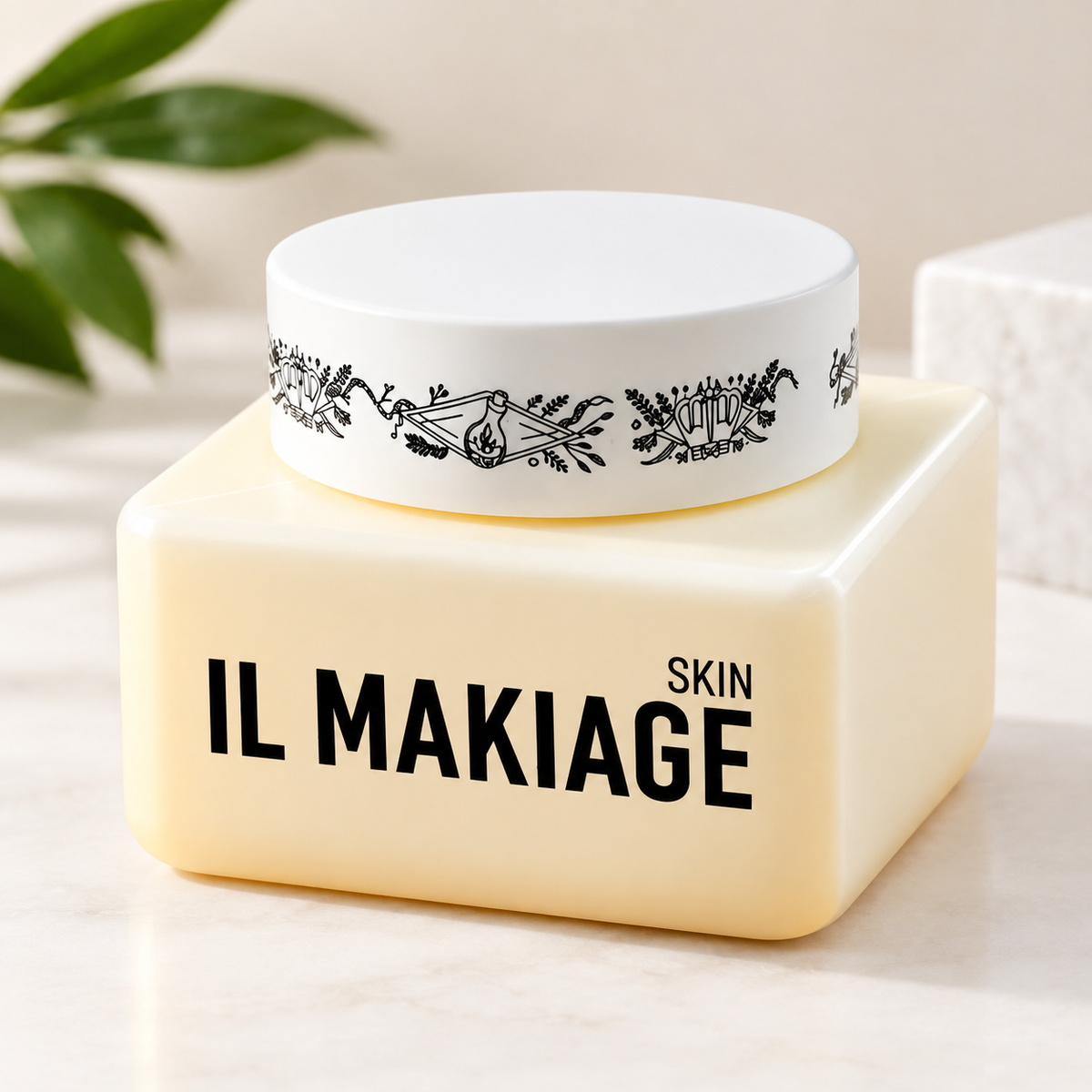 IL MAKIAGE SKIN cream in a square ivory jar with a white botanical-patterned lid, displayed on a light beige surface with soft greenery in the background.