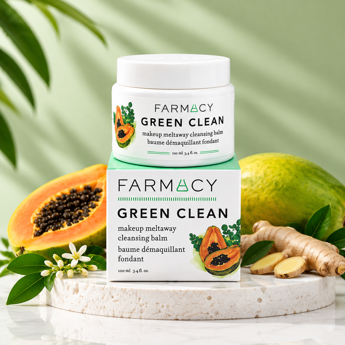 A Farmacy Green Clean makeup meltaway cleansing balm jar and box displayed on a marble surface, surrounded by green leaves, okra-like vegetables, and a halved papaya, with lush plants in the background.