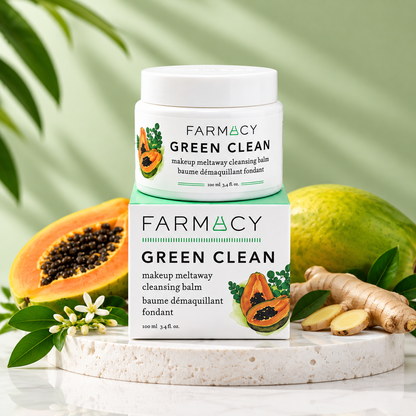 A Farmacy Green Clean makeup meltaway cleansing balm jar and box displayed on a marble surface, surrounded by green leaves, okra-like vegetables, and a halved papaya, with lush plants in the background.
